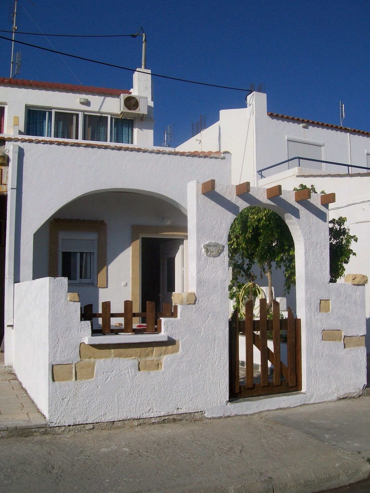 The exterior of a whitewashed building features a traditional archway with wooden accents and a small fence, leading to a welcoming entrance. Sunlight highlights the structural details against a clear blue sky, emphasizing the charming simplicity of the design.