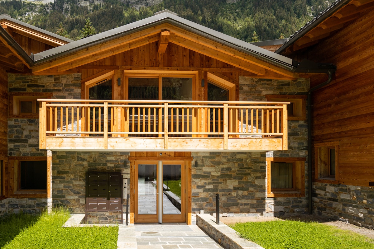 The chalet-style building features a wooden facade with a sloped roof, large windows, and a balcony. The entrance is framed by stone walls and a wooden canopy, providing a welcoming atmosphere. A pathway leads through green grass towards the door.