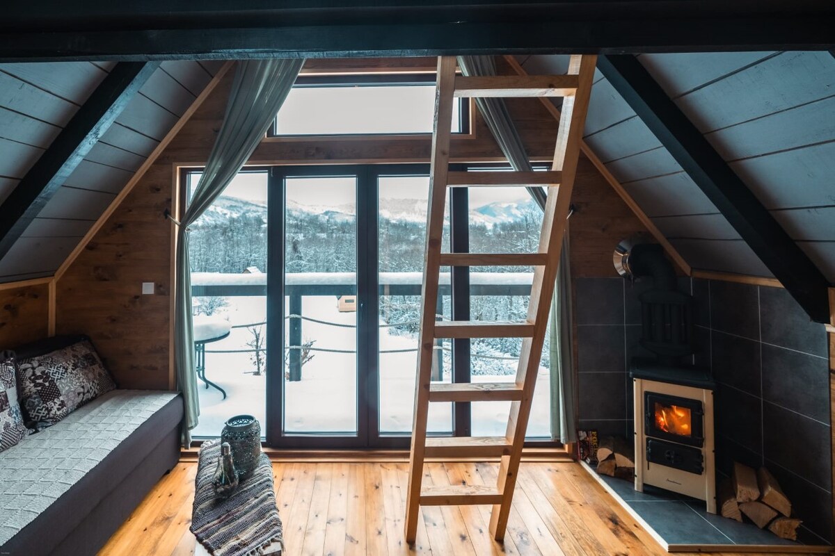 A cozy living space with an inviting wood stove is featured next to large windows offering panoramic winter views. A ladder leads to an upper sleeping area, while a comfortable sofa and decorative items add warmth to the atmosphere.