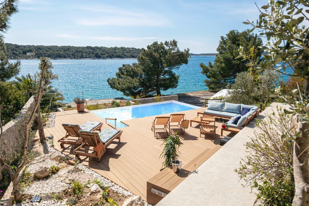 An outdoor space showcases a heated pool surrounded by wooden decking. Sun loungers and a seating area are positioned to offer views of the serene waters and lush trees in the background. The area is enhanced by well-maintained greenery and inviting natural light.