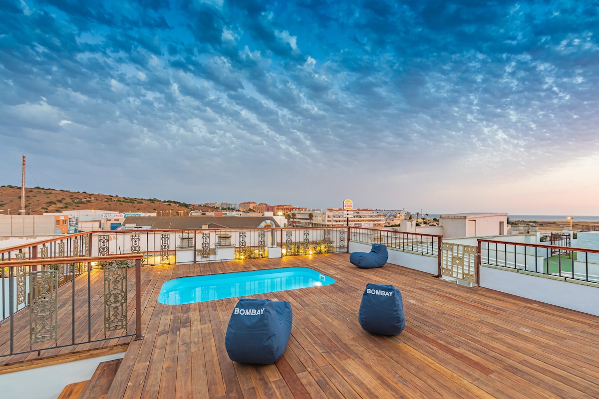 An expansive terrace features a turquoise pool surrounded by wooden decking and casual seating. Soft, blue bean bags provide comfortable lounging options. The view extends towards the horizon, with a mix of natural and urban scenery under a dramatic sky.