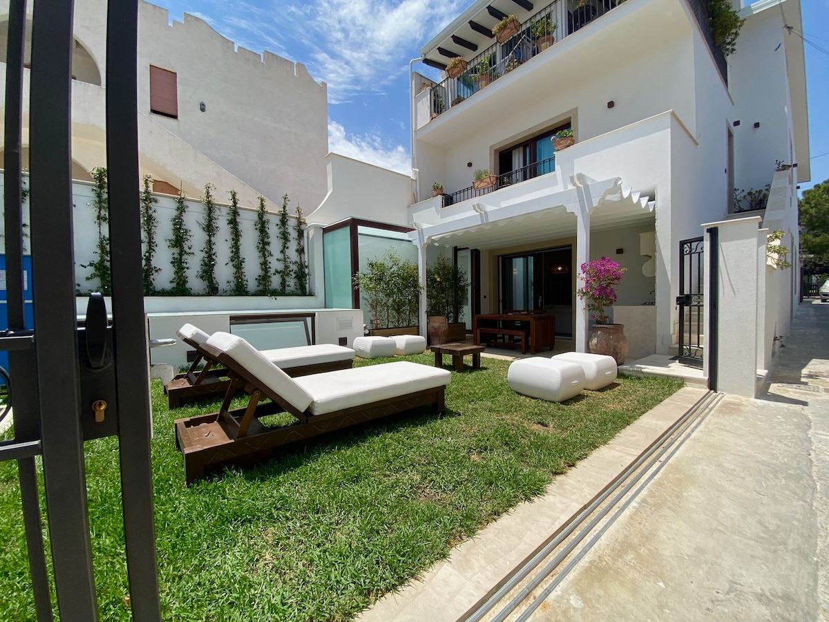 A spacious outdoor area is featured, showcasing a well-maintained lawn. Multiple lounge chairs are positioned for relaxation, surrounded by flowering plants. The white facade of the building and large glass windows provide a bright backdrop, enhancing the inviting atmosphere of the terrace.