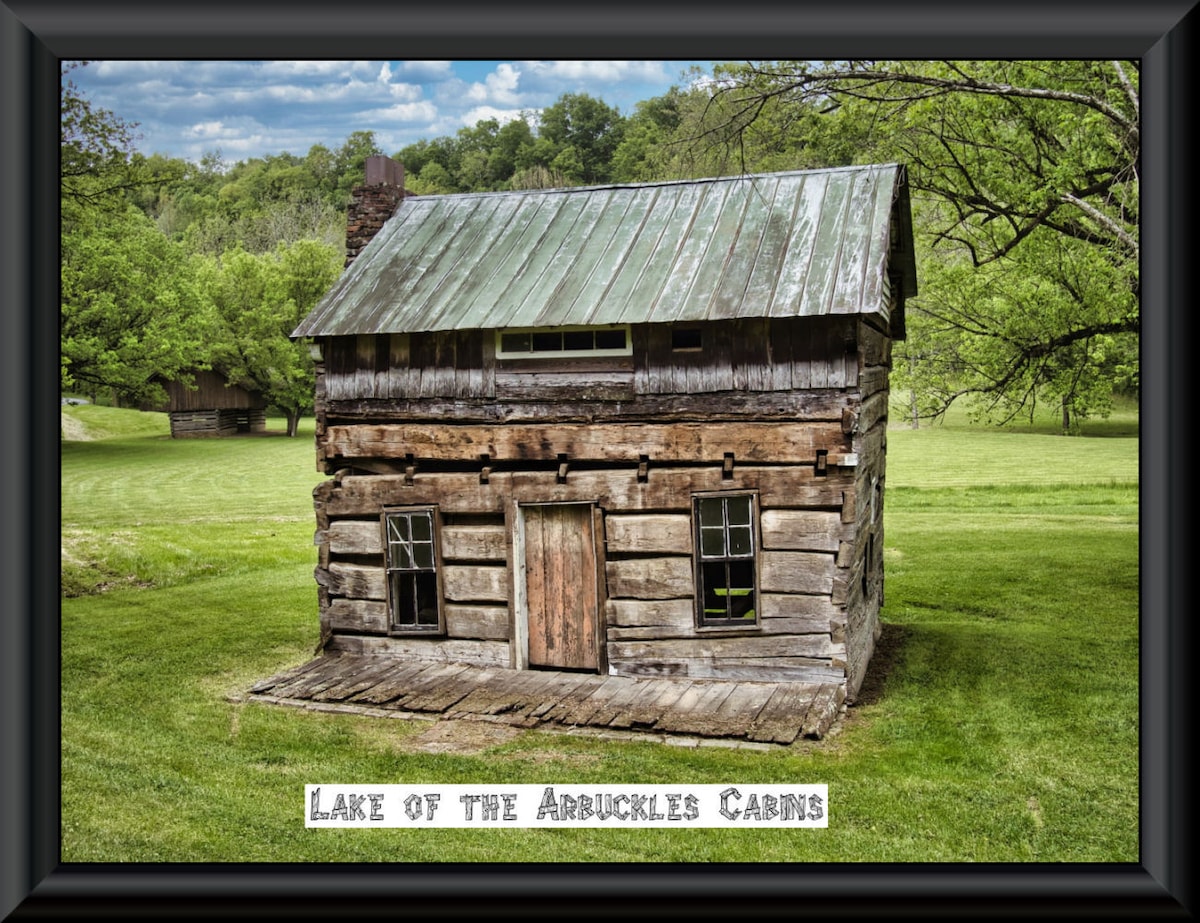 An aged log cabin with a sloped metal roof is surrounded by green grass and trees. The structure features distinct wooden planks and two small windows on each side. A wooden door with a rustic appearance is centered at the front, inviting exploration.