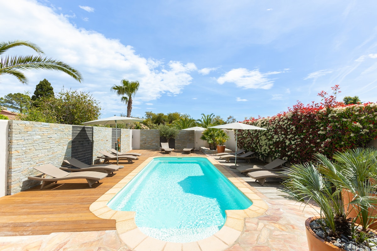 A shared outdoor pool is surrounded by a wooden deck with several loungers beneath large umbrellas. Lush greenery and flowering plants enhance the private ambiance, while stone walls provide a sense of seclusion. The inviting pool is illuminated by natural sunlight under a blue sky.