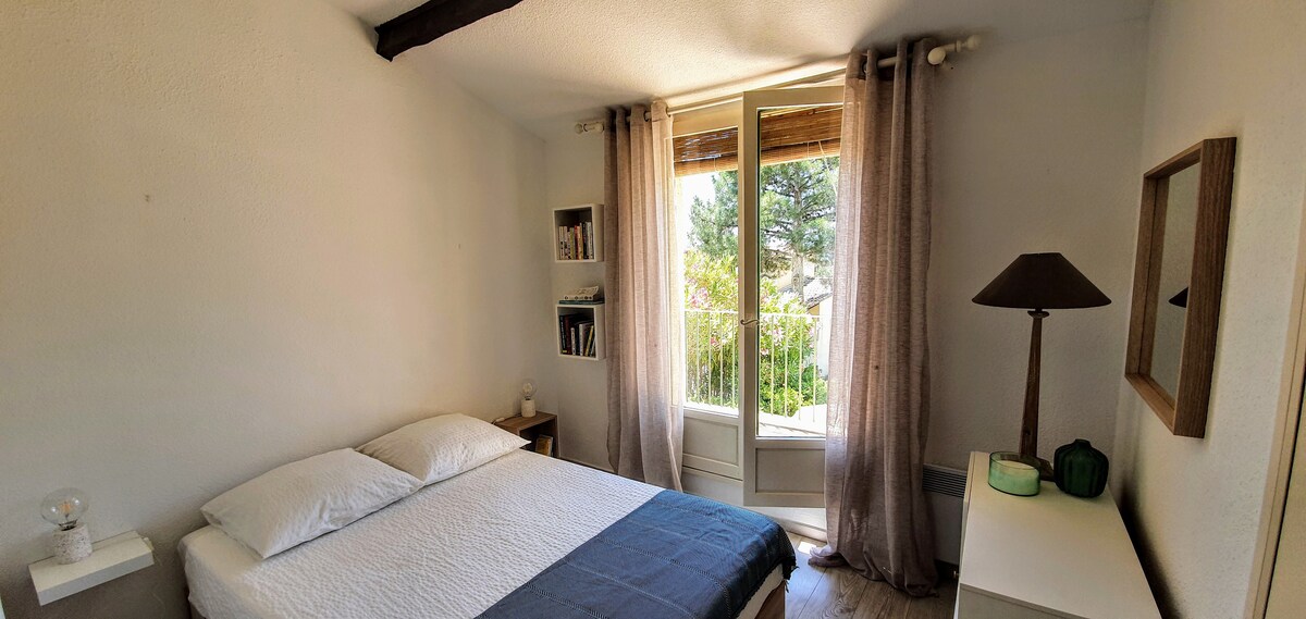 A bright bedroom features a spacious bed dressed in soft linens. Natural light filters through large windows covered with sheer curtains, highlighting a bedside lamp and a decorative mirror. A small bookshelf is visible, adding a touch of comfort to the space.