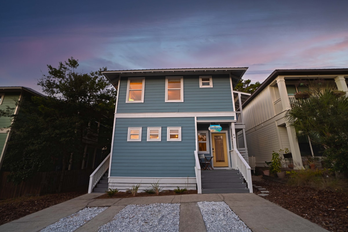 A two-story beach house with a modern, blue exterior is shown at dusk. The entryway is highlighted by a welcoming door and is flanked by narrow windows, while steps lead up to the elevated porch. Gravel pathways and landscaped plantings enhance the front yard.