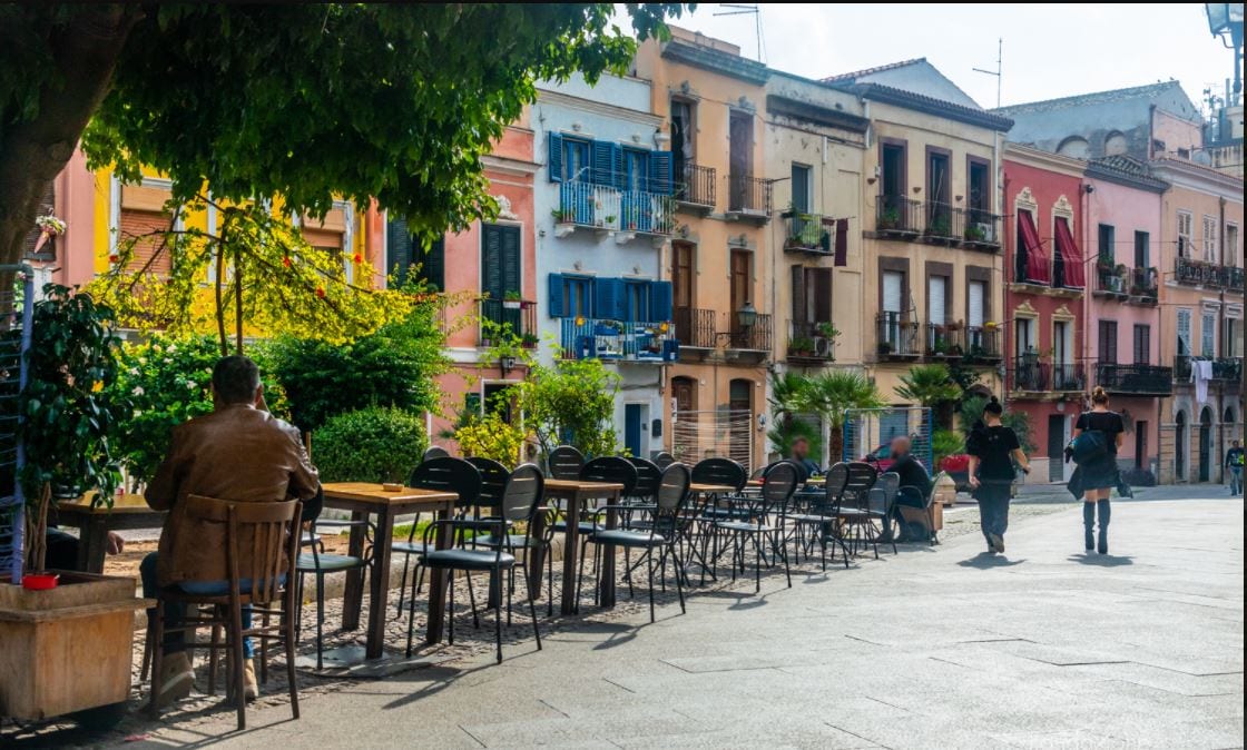 A lively street scene is depicted, featuring an outdoor seating area with wooden tables and black chairs. Colorful buildings with balconies and greenery line the street, creating a welcoming atmosphere. People are seen engaged in conversation, adding to the vibrant community feel.