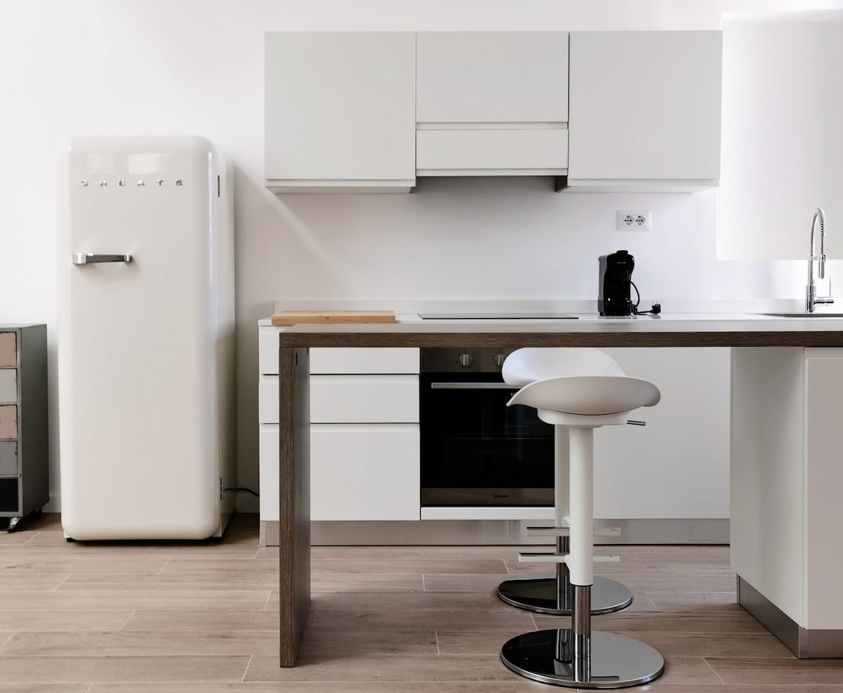 A modern kitchen area features sleek white cabinetry and a retro-inspired refrigerator. A wooden island with a light-colored stool provides a casual dining space. The kitchen is well-lit, with appliances neatly integrated and surfaces kept clear.