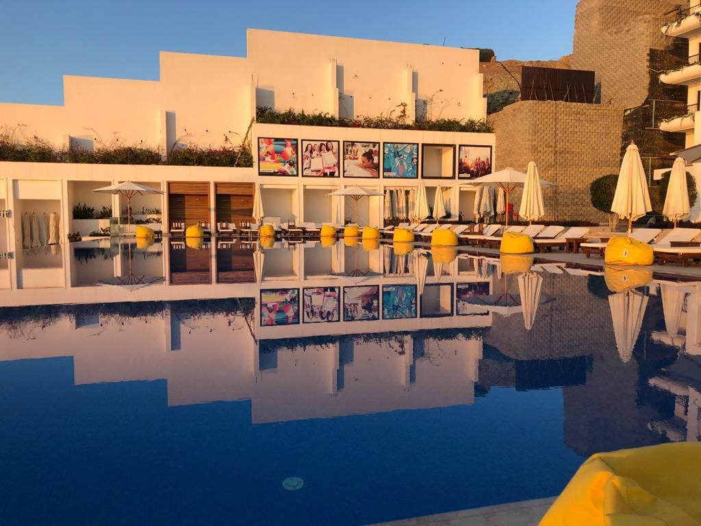 A serene swimming pool reflects the surrounding architecture and vibrant art displays. Sun loungers with yellow cushions are arranged along the poolside, accompanied by white umbrellas providing shade. The minimalist design of the building enhances the tranquil atmosphere.