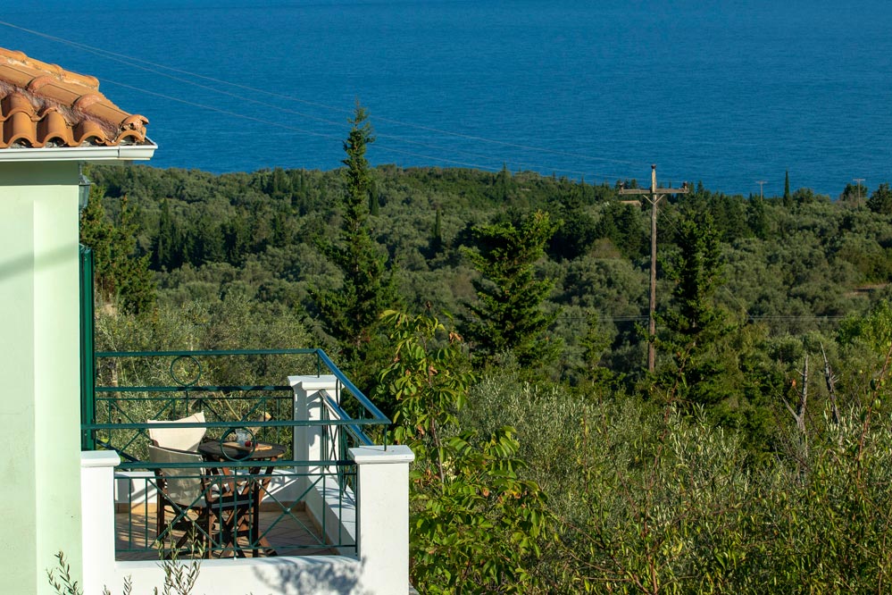 A balcony is shown overlooking lush greenery and the sea. A small table with chairs is positioned on the balcony, providing a serene spot to enjoy the views. The terracotta roof adds a touch of rustic charm to the setting.
