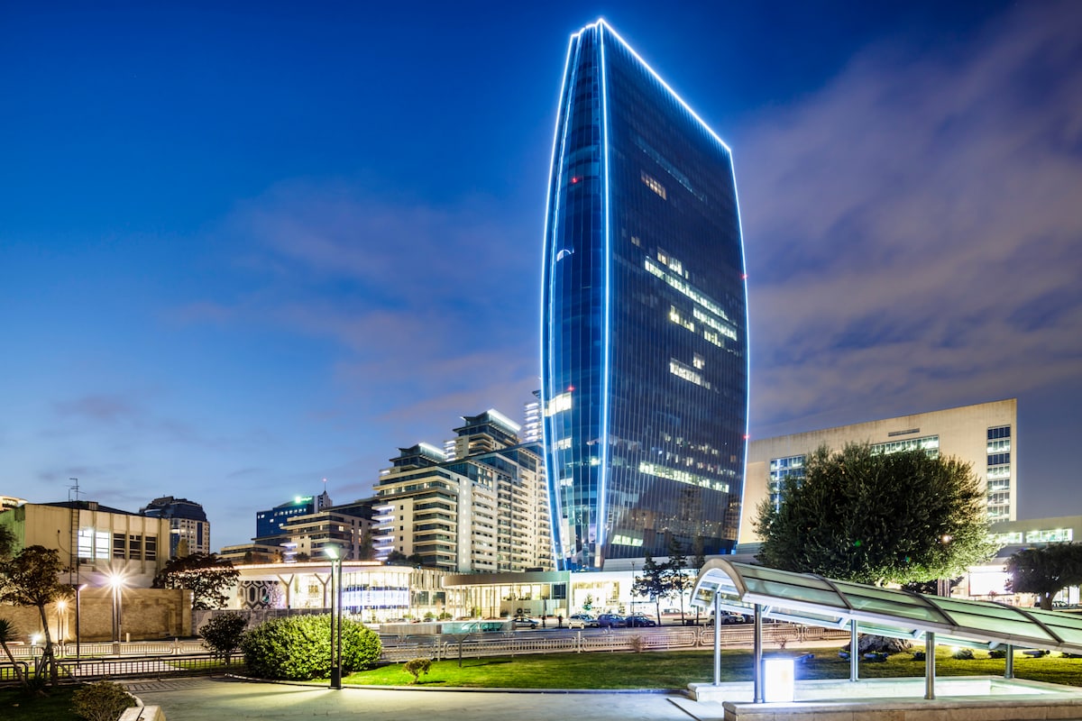 The towering luxury residence is illuminated with blue lighting, contrasting against the evening sky. Surrounding buildings showcase modern architecture, with greenery defining the lower landscaping. The vibrant city circuit is visible in the foreground, emphasizing the location's urban setting.