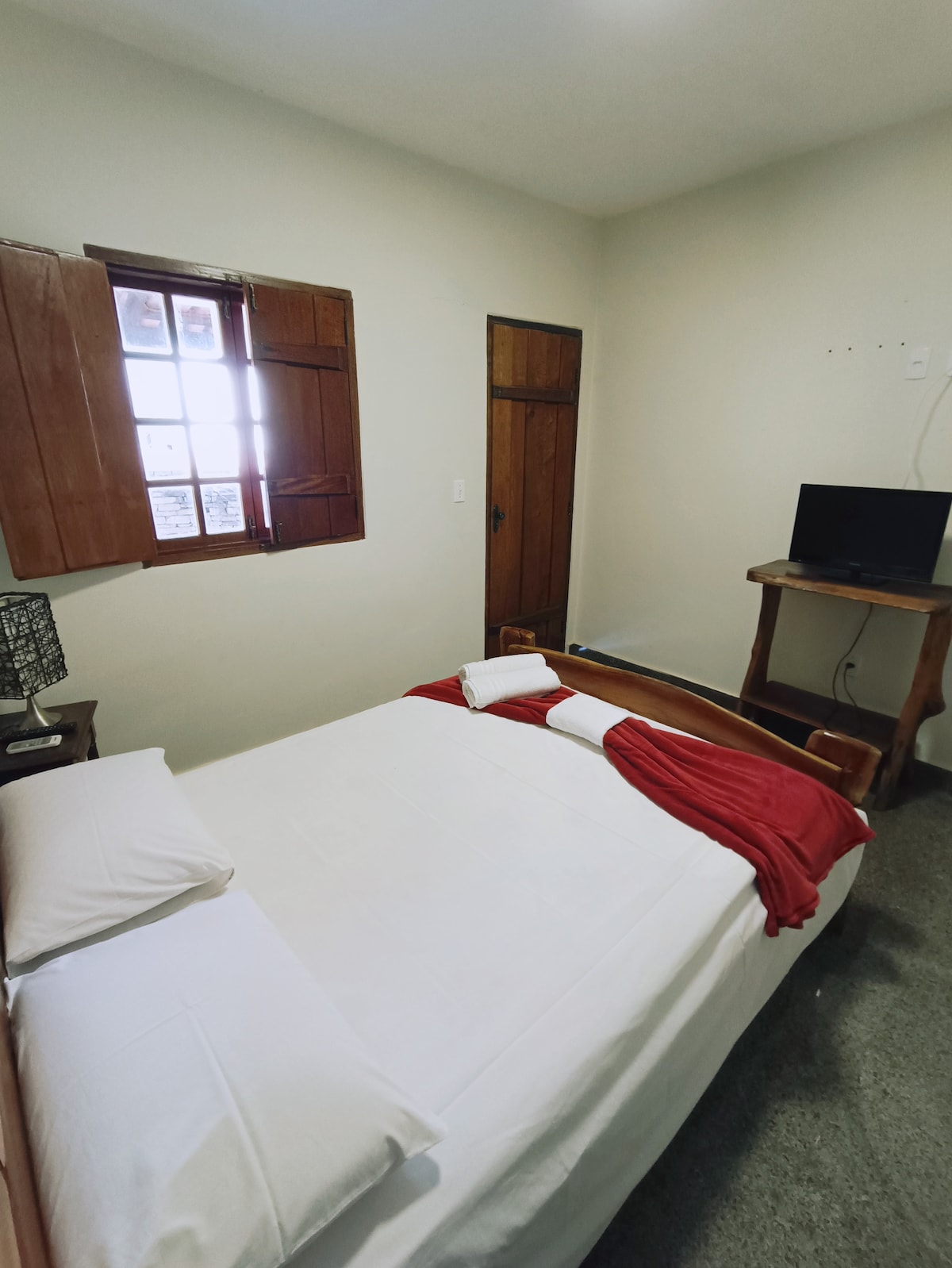 A cozy bedroom features a spacious bed adorned with white linens and a red throw. Natural light enters through a window with wooden shutters, complementing the neutral-colored walls. A small television is positioned on a wooden stand, enhancing the functional space.