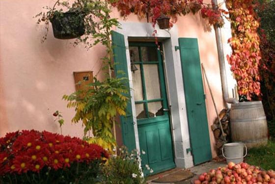 An inviting exterior features a green door framed by vibrant foliage and a seasonal display of red flowers. A wooden barrel and a collection of apples are visible on the ground, contributing to the charming rural ambiance.