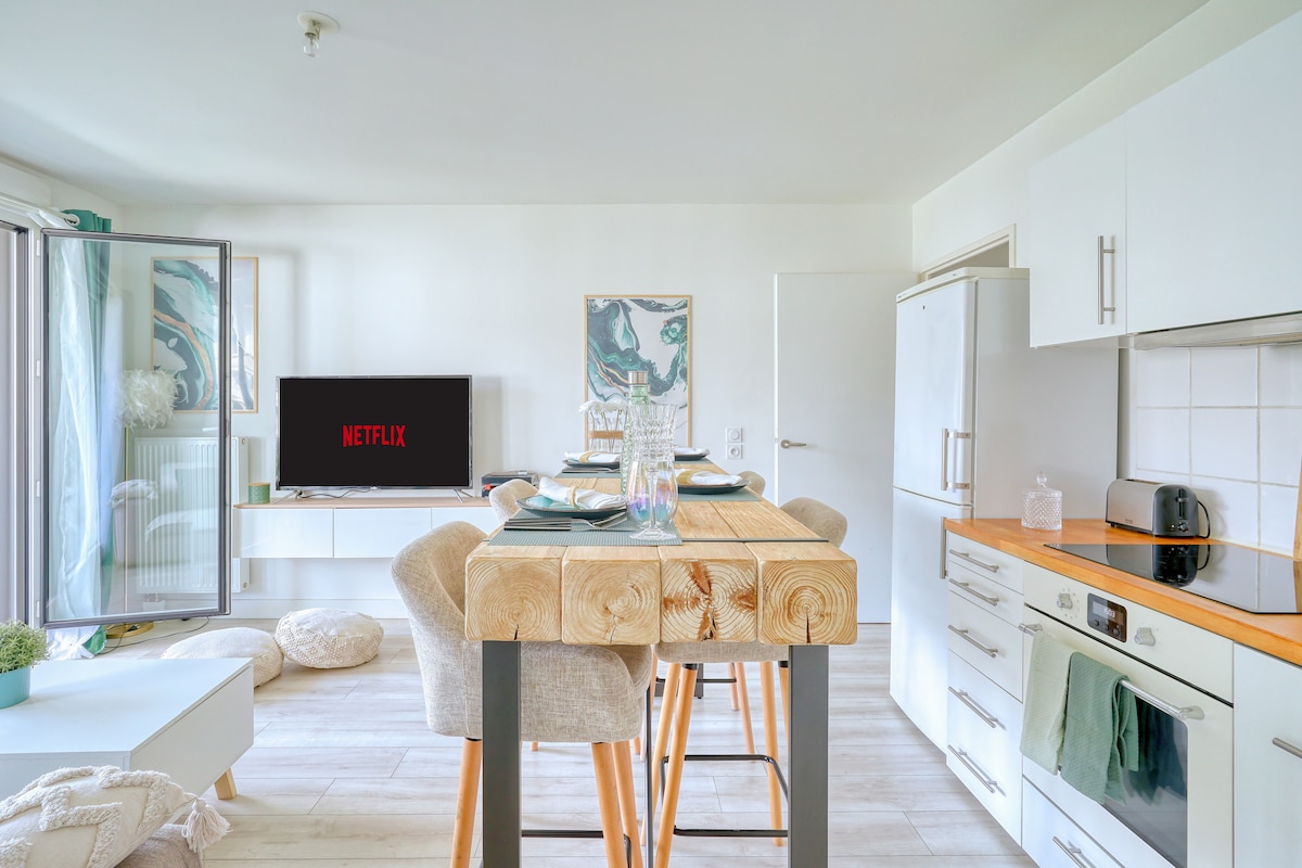 A bright living area is highlighted by a wooden dining table with high stools. A flat-screen TV displays the Netflix logo, while a modern, fully equipped kitchen is visible with sleek white cabinetry and appliances. Natural light enters through a sliding glass door leading to a terrace.