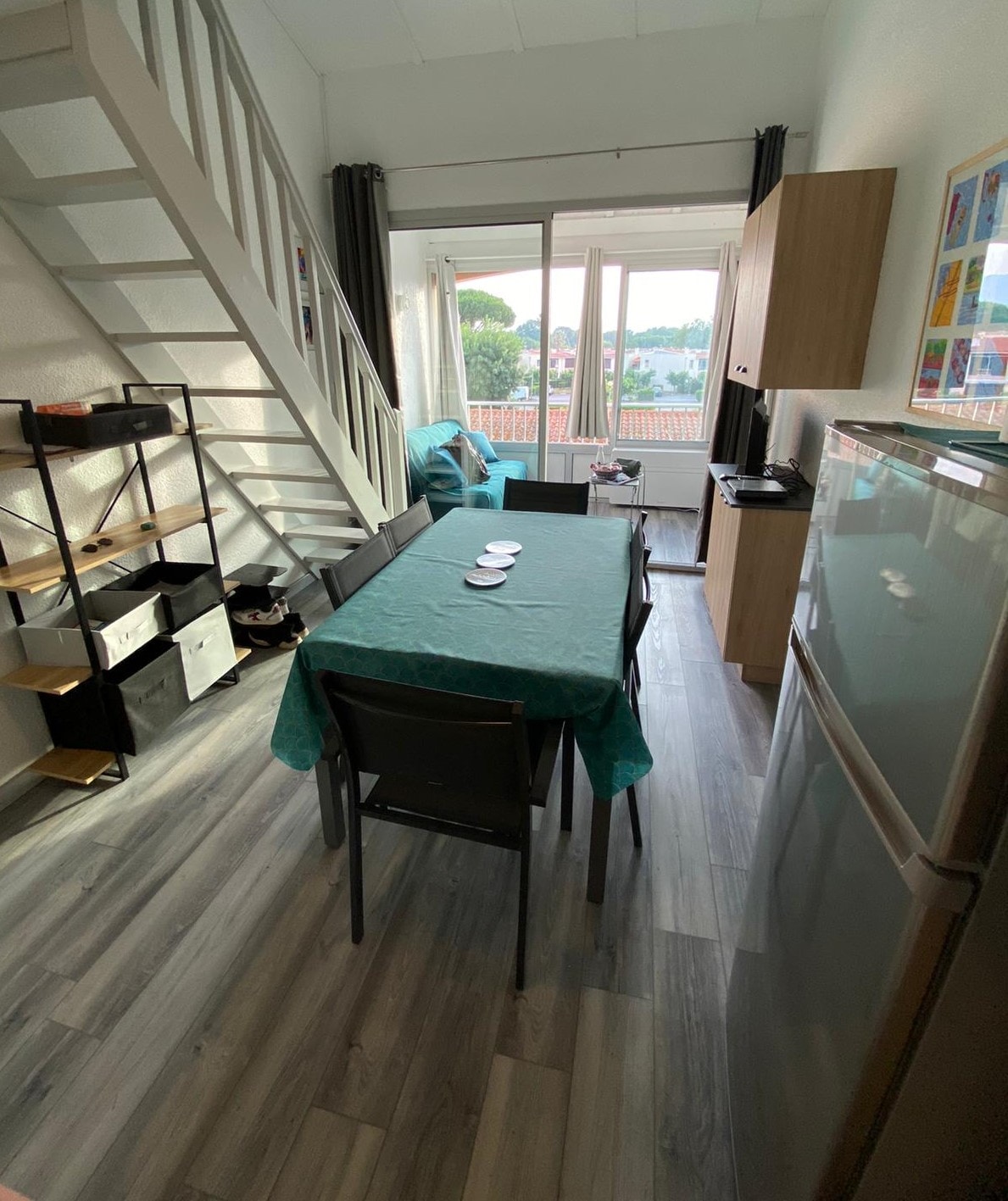 A bright dining area features a rectangular table covered with a turquoise tablecloth, surrounded by chairs. Natural light filters through the large window, illuminating the space. A staircase leads to an upper level, and a refrigerator is positioned nearby, along with a storage unit displaying various organizers.