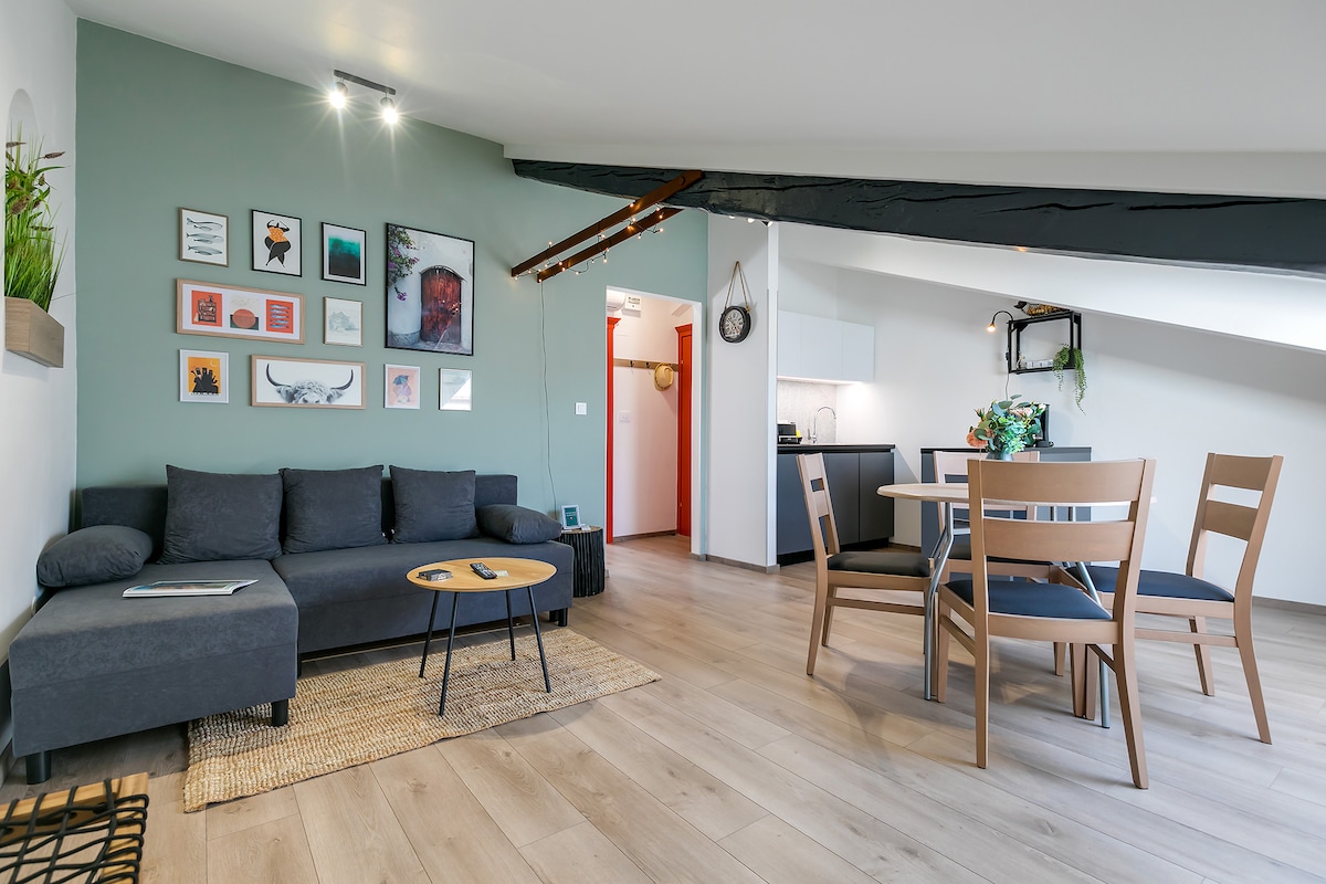A modern living space features a dark sofa accompanied by a small round table and four wooden chairs. A gallery wall displays various framed artworks. The open layout includes a kitchenette visible in the background, with natural light streaming through a roof window.