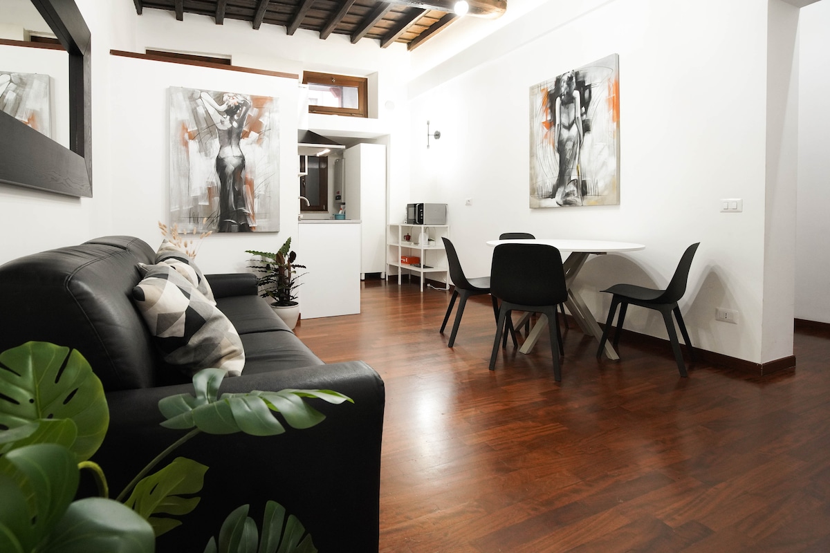 The living area features a modern black sofa paired with decorative cushions. A dining table with sleek chairs is situated nearby. Light wooden beams are visible on the ceiling, while abstract artwork adorns the walls. Lush green plants add a touch of nature to the space.