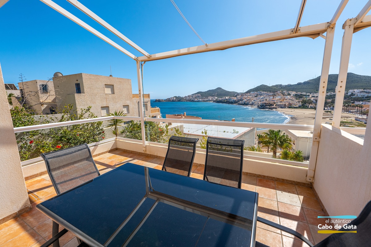 A spacious terrace is shown with a table and chairs, offering panoramic views of the coastline and distant hills. The bright blue sky complements the tranquil sea, creating a serene outdoor space perfect for relaxation.