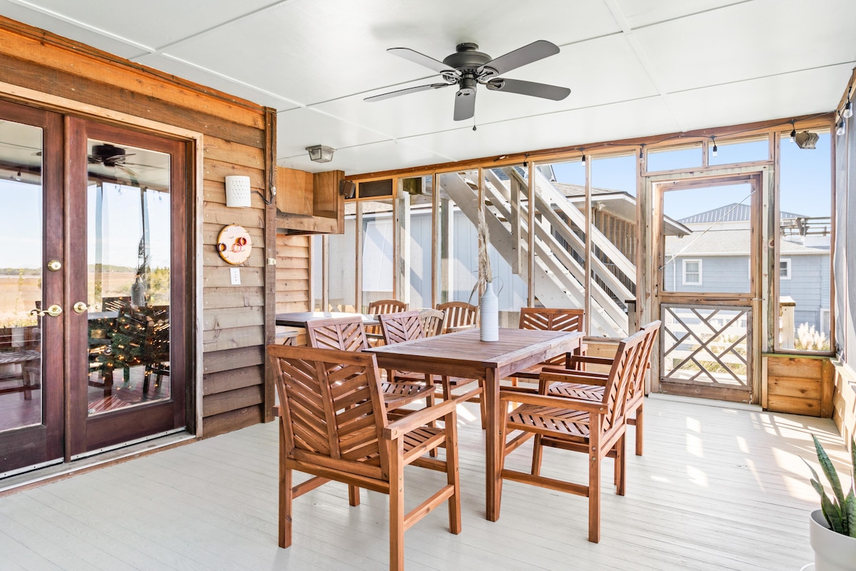 A spacious screened-in porch features a large wooden dining table surrounded by matching chairs. Natural light fills the area, highlighting the wooden walls and ceiling fan. Double doors lead to the interior spaces, enhancing the open atmosphere.