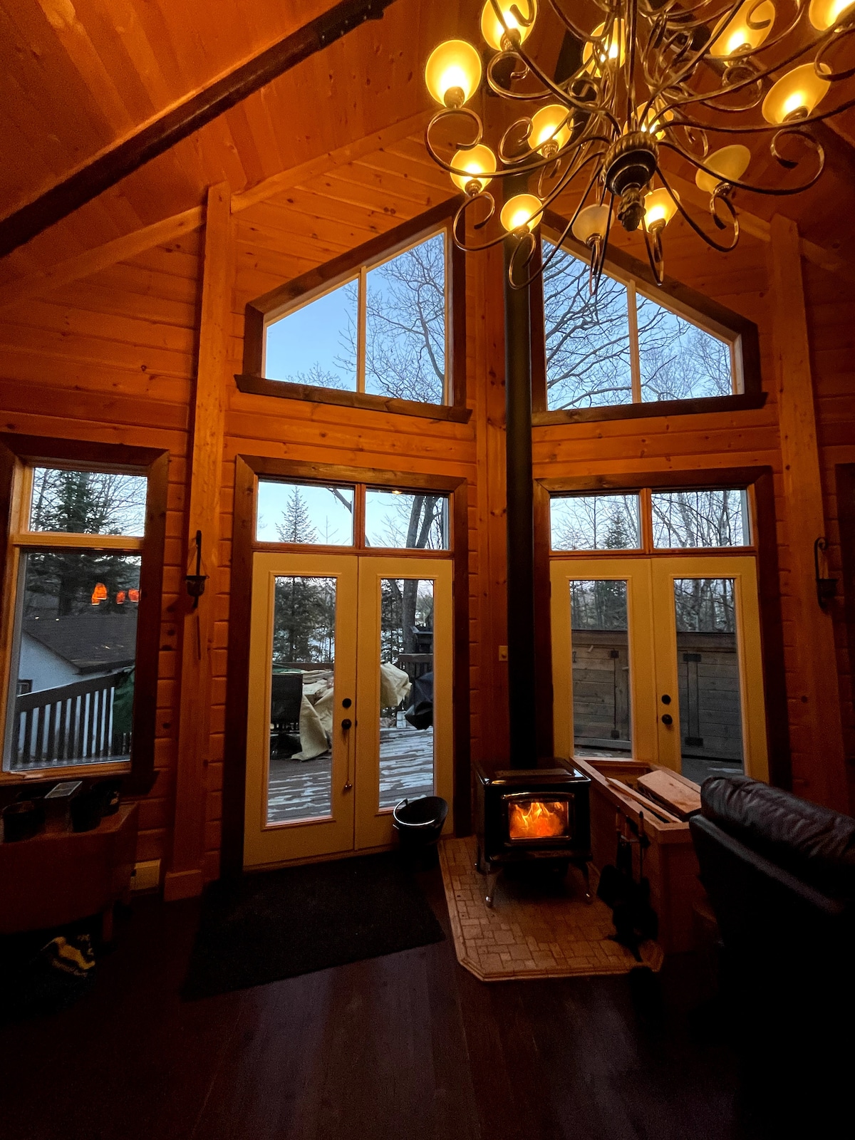A cozy living area features large windows allowing natural light to fill the space, highlighting the wooden walls and ceiling. A gas stove provides warmth, with a rustic coffee table positioned in front of a comfortable sofa. The deck is visible through the glass doors.