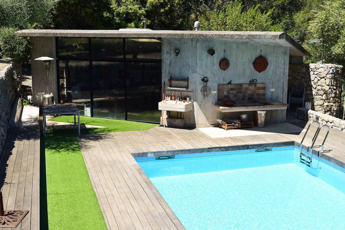 A modern pool area features a refreshing blue pool surrounded by wooden decking and artificial grass. Adjacent, a concrete pool house displays a barbecue area and rustic decor, with large glass doors offering a view into the interior space. Lush greenery frames the scene.