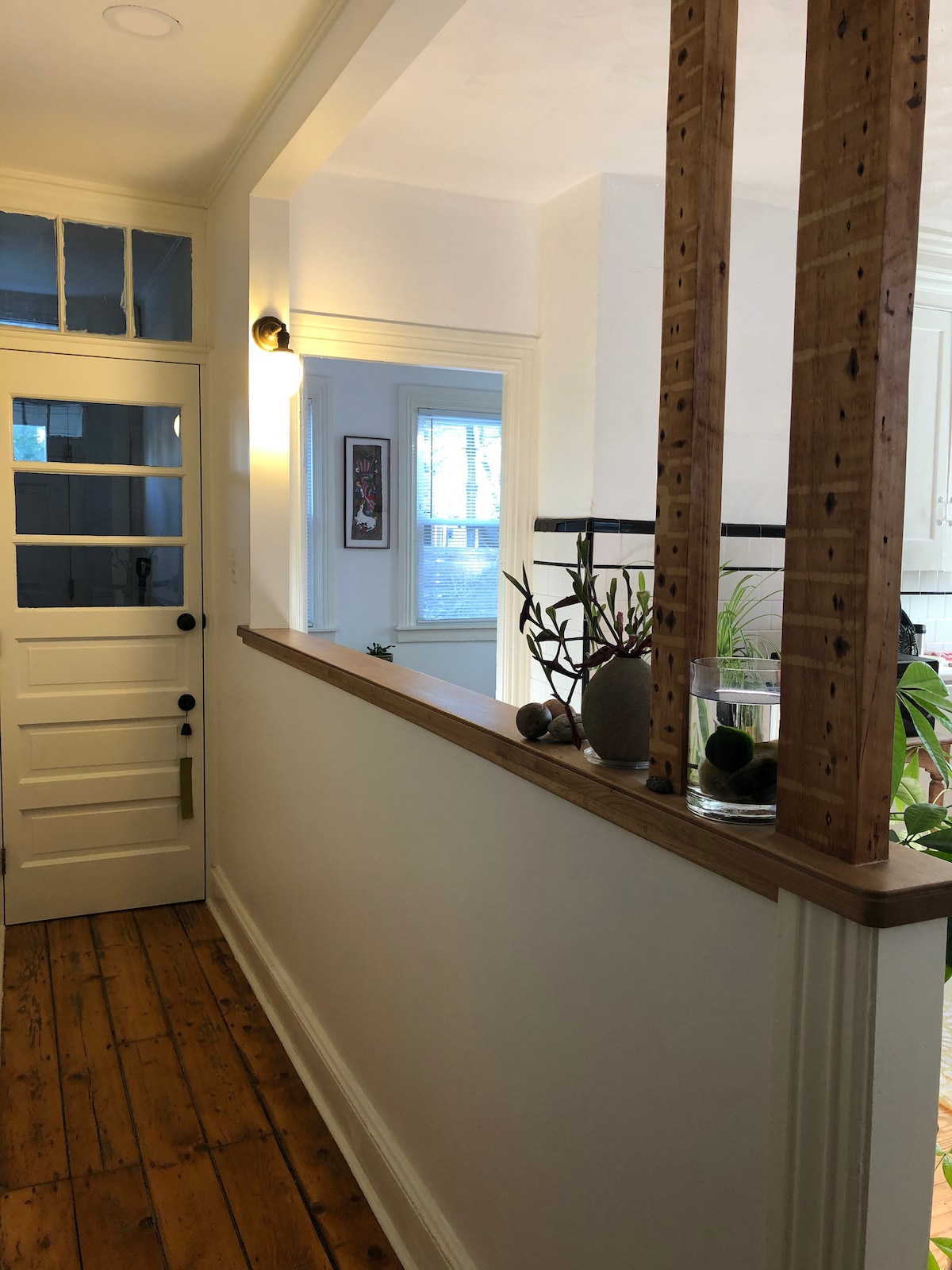 A well-lit hallway features natural wood flooring and white walls, creating an open feel. A glass vase with decorative stones sits on a ledge, and a doorway leads to a room beyond. Potted plants add a touch of greenery to the space.