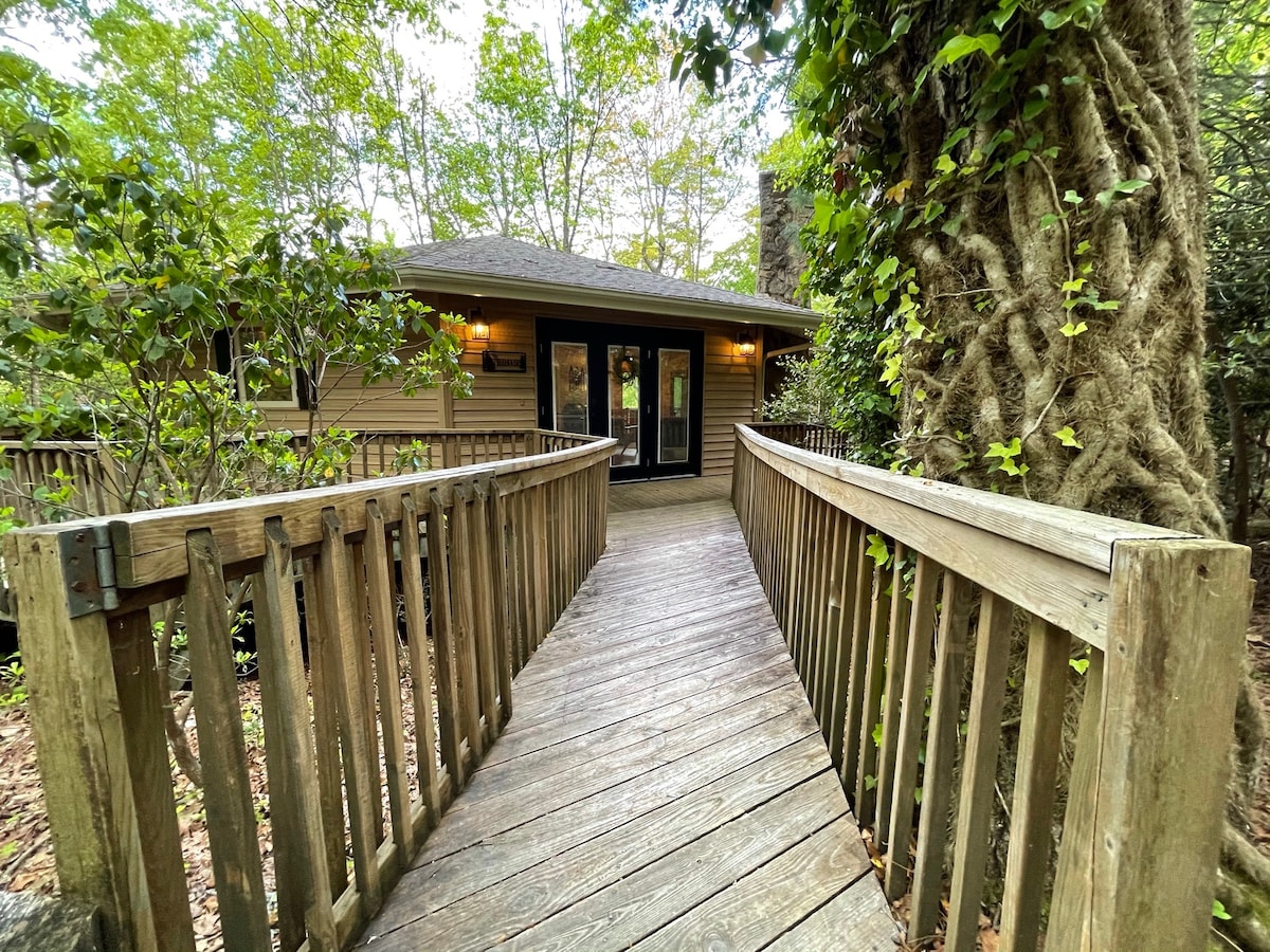 A wooden walkway leads to the entrance of the cabin, framed by lush greenery and trees. The doorway features large glass panels flanked by warm outdoor lighting, creating an inviting entrance into the nature-surrounded space.