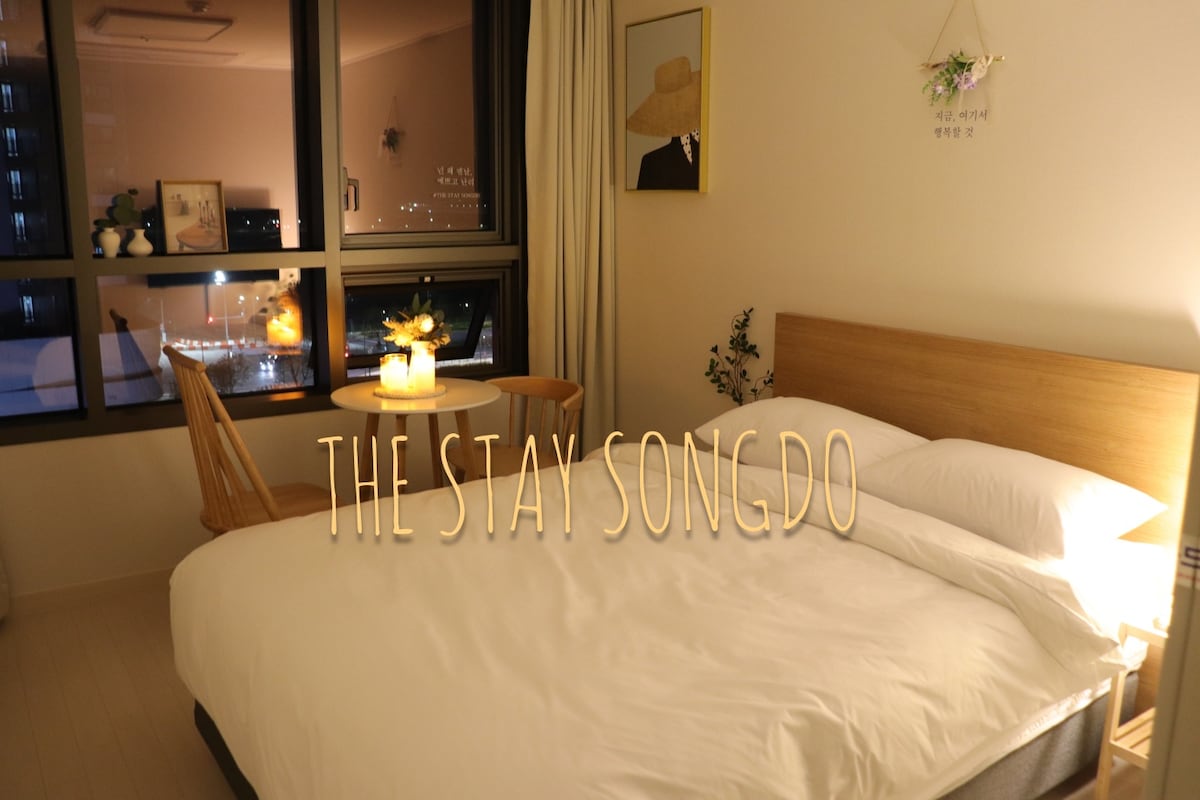 A cozy bedroom features a spacious bed with hotel-style bedding. A small round table with two chairs is positioned near a large window, offering a view of the city lights. Soft ambient lighting enhances the inviting atmosphere.