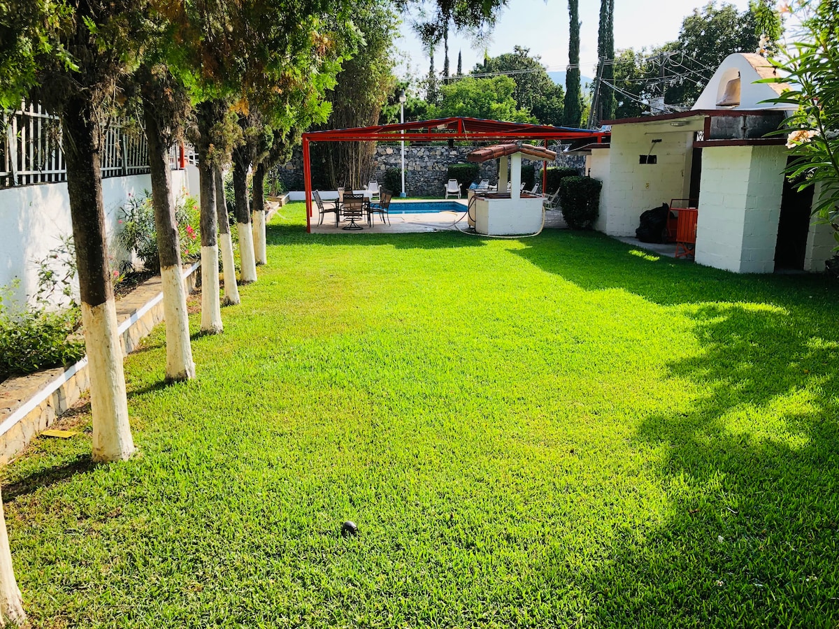 A sunlit grassy area is framed by neatly trimmed trees, leading toward a shaded patio with a roof. The inviting pool area can be seen in the background, surrounded by lush greenery, providing a serene outdoor setting for relaxation.
