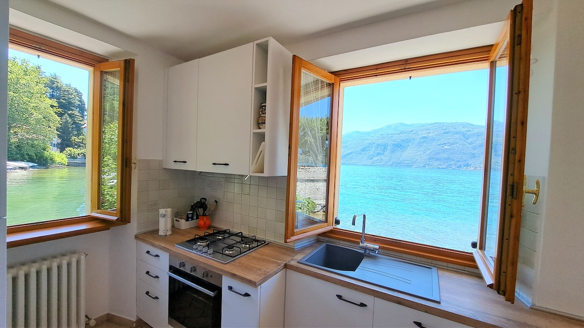 A bright kitchen space features modern cabinetry and a windowed view of the lake. Natural light floods the area through two open windows, enhancing the serene atmosphere. Kitchen amenities include a gas stove, sink, and countertop with a clean, minimalist design.