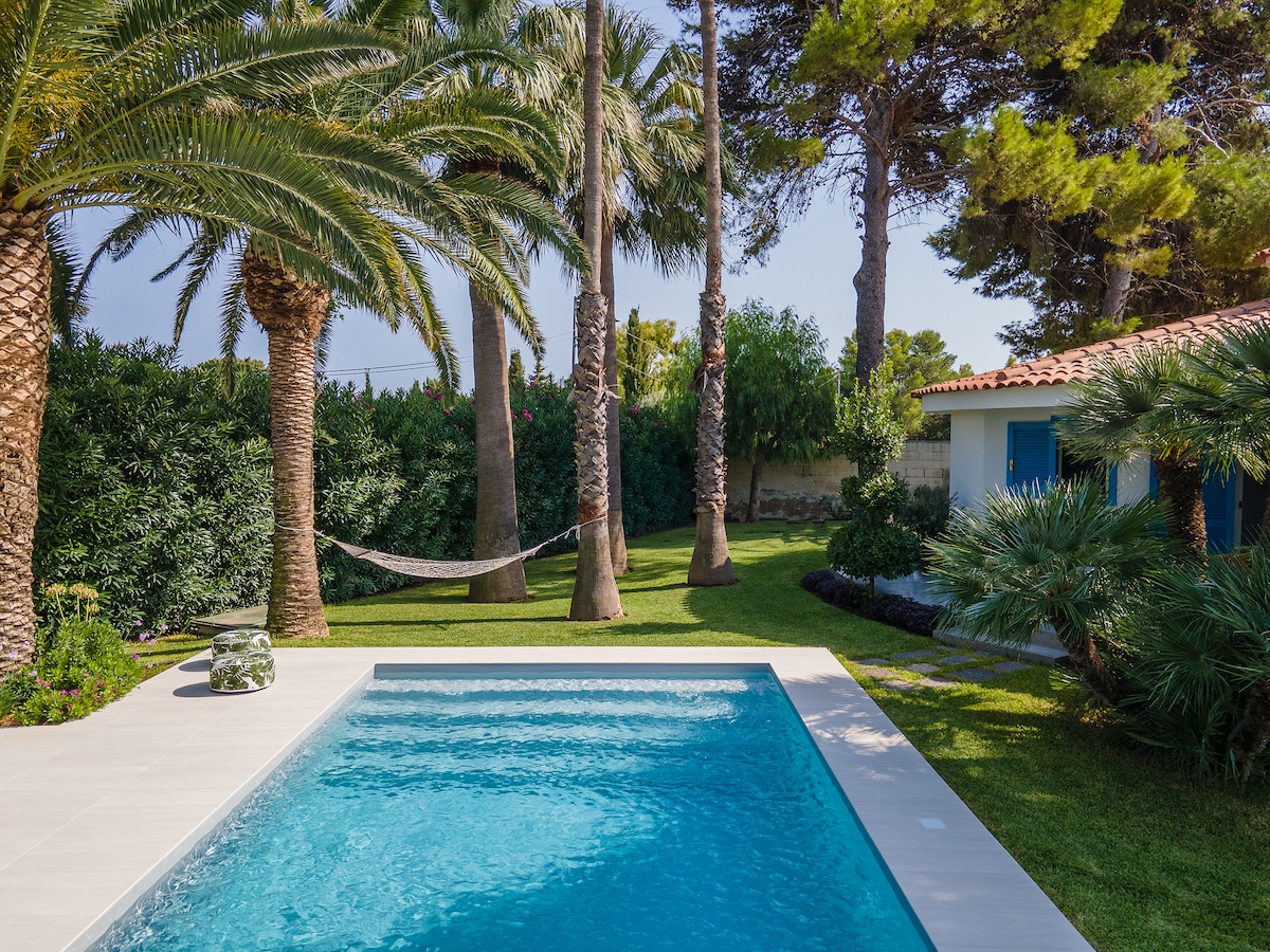A private swimming pool is surrounded by lush greenery and palm trees, offering a serene atmosphere. A hammock is visible between two trees in the background, complementing the tranquil garden setting. The pool area is designed with light-colored tiles, enhancing the inviting outdoor space.