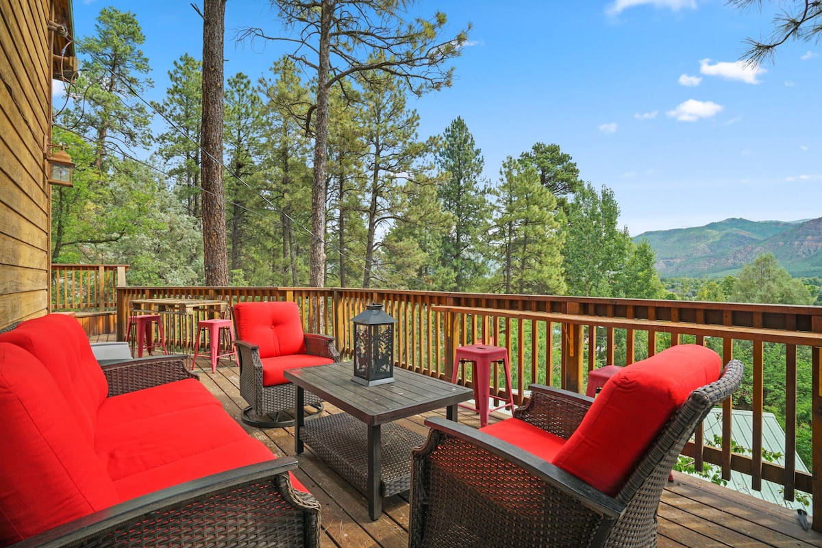 A spacious deck features a seating area with red cushions on woven chairs, complemented by a coffee table. Surrounded by trees, the area provides expansive views of the mountains and valley, creating an inviting setting for relaxation.