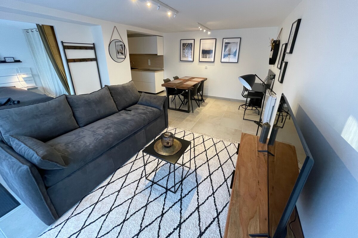 A spacious living area is furnished with a gray sofa and a decorative coffee table atop a geometric rug. A dining table with black chairs is visible, alongside a modern kitchenette and framed artwork on the wall. Natural light fills the room from a nearby window.