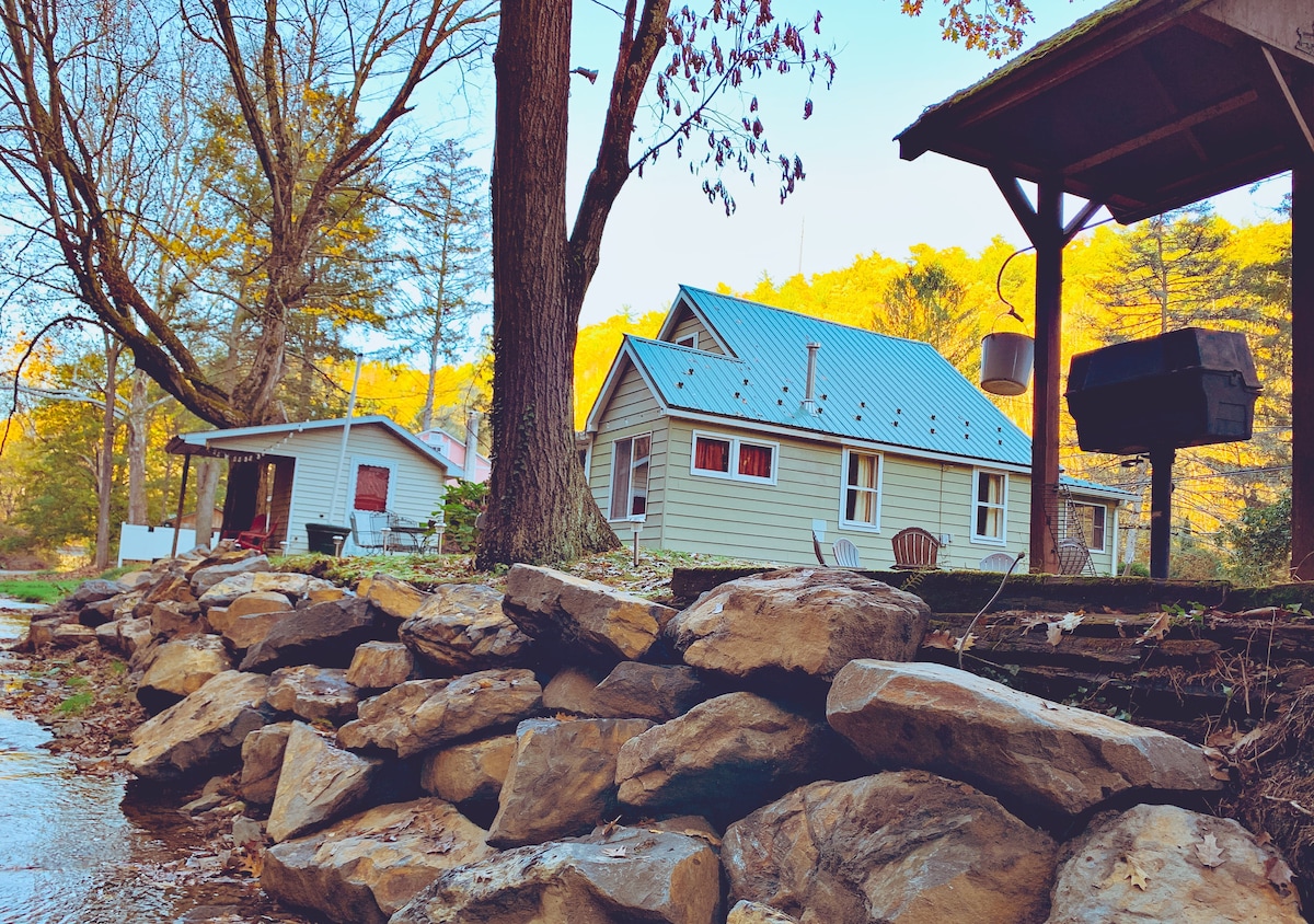 A serene view of Coaster Creek Cottage sits alongside a gently flowing creek, framed by lush trees. A spacious deck features a charcoal grill, while the cottage's exterior showcases a light color palette with red accents and large windows. Natural stone creates a rustic border along the water.