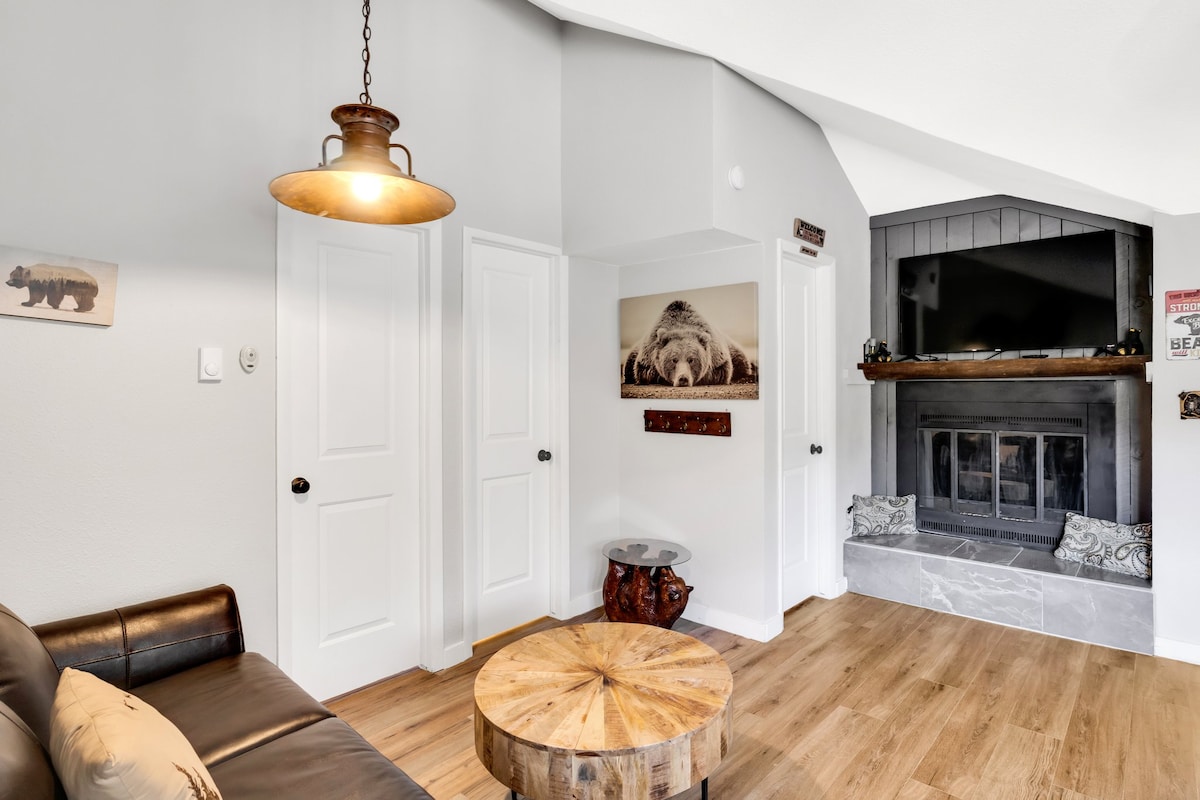 A cozy living area features a dark leather sofa and a round wooden coffee table. A stone fireplace sits beneath a mounted television, with decorative accents placed on the mantel. Soft light is provided by an industrial-style pendant light hanging from the ceiling.