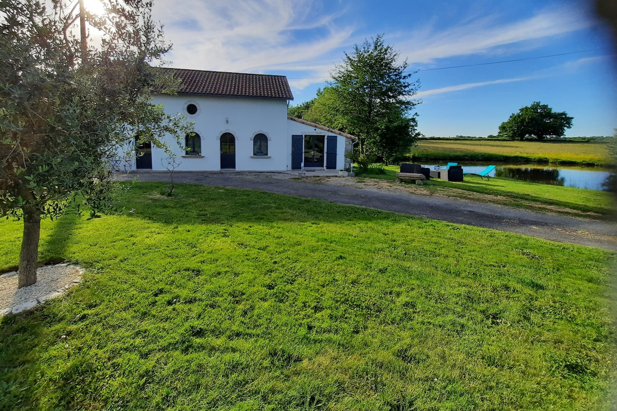 The exterior of the renovated gîte is shown, featuring a white facade with arched windows. A grassy area surrounds the house, with a tree on the left and a pond visible in the background. Outdoor seating is positioned near the pond, providing a serene setting.