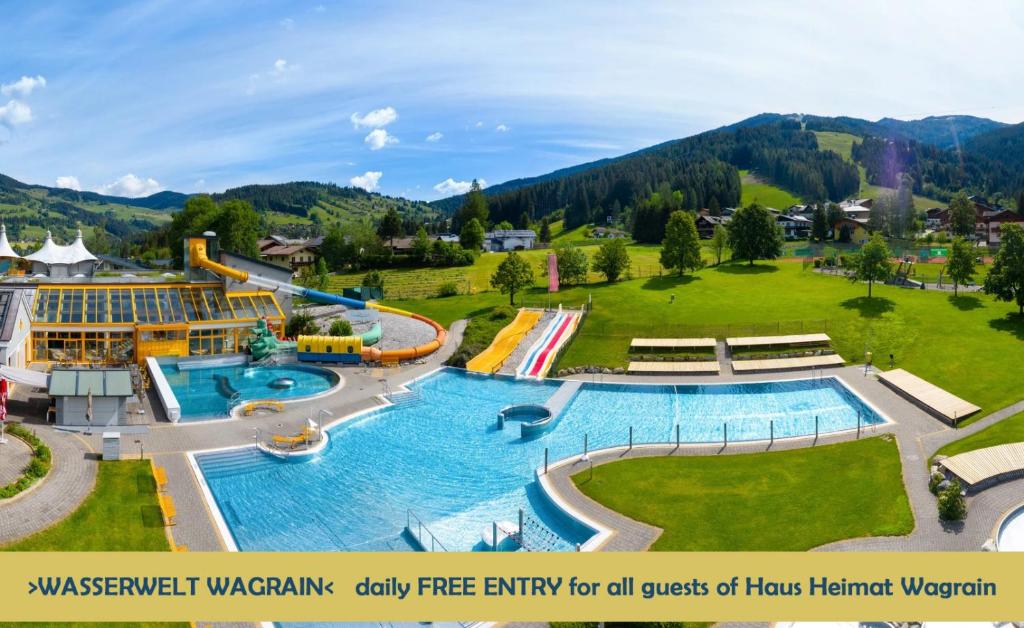 An expansive outdoor water park is showcased, featuring multiple swimming pools and colorful water slides. Lush green hills serve as a backdrop, while a spacious lawn is visible, providing ample seating areas. Promotional text indicates daily free entry for guests of Haus Heimat Wagrain.