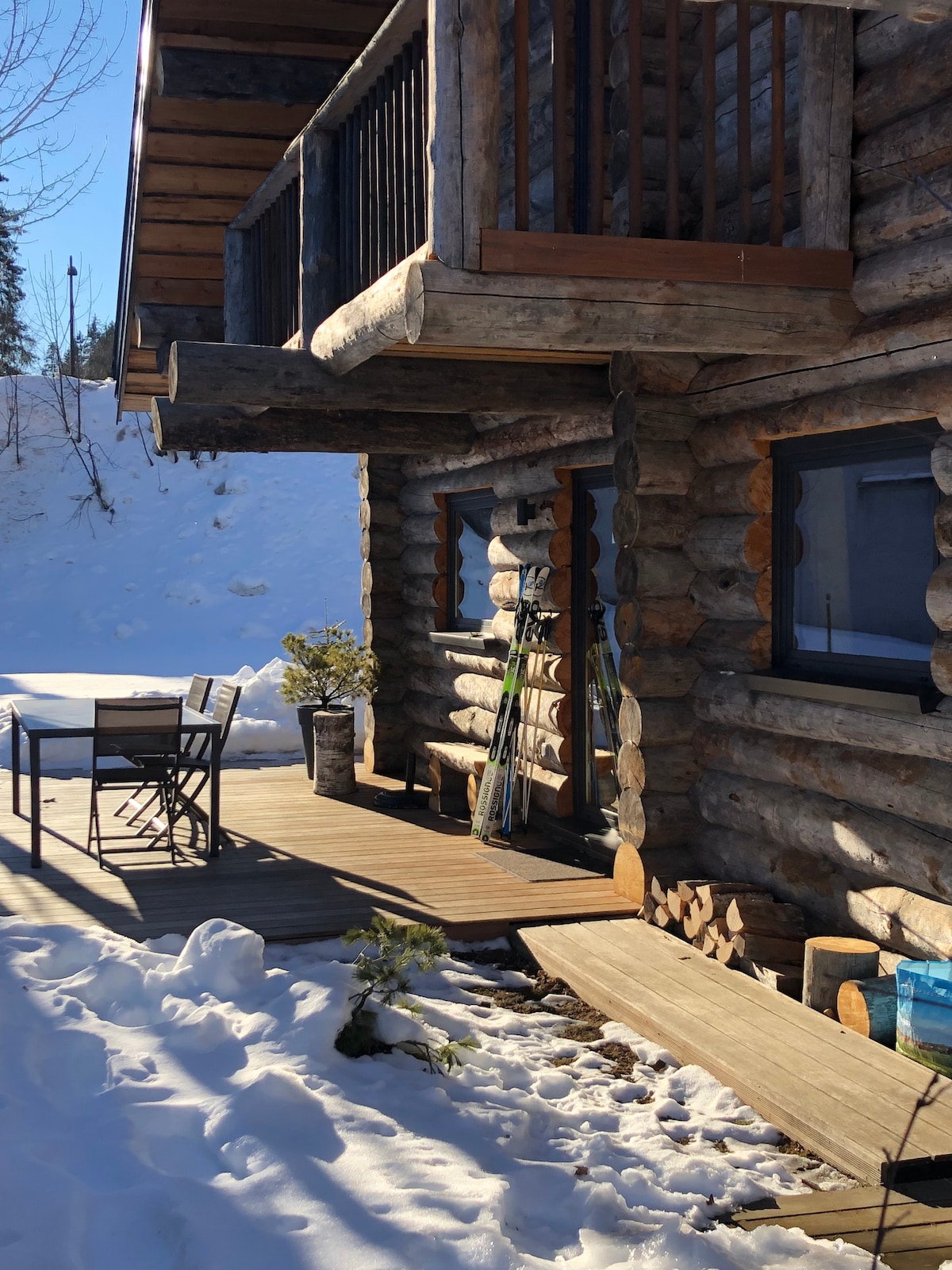 The exterior of a log cabin is presented, featuring a wooden deck equipped with a dining table and chairs. Snow blankets the ground, and skis are leaned against the wall. Sunlight casts shadows, highlighting the texture of the logs and the surrounding winter landscape.