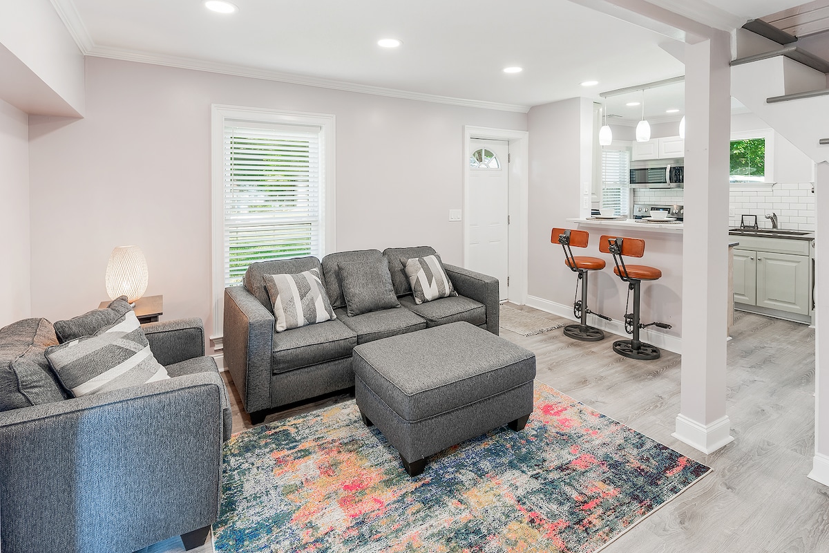 A spacious living area features comfortable gray sofas adorned with pillows, centered around a stylish coffee table. Natural light fills the room through the window, while sleek bar seating for two is positioned at the kitchen counter in the background.