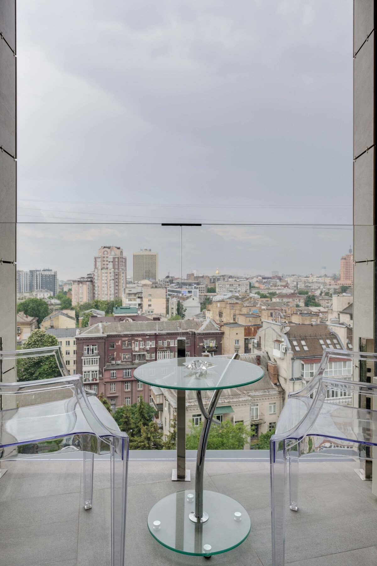 A balcony features a glass table and two transparent chairs, offering a view of the cityscape. A mix of modern and historical buildings is visible, with greenery interspersed throughout. The sky is overcast, contributing to a serene atmosphere.