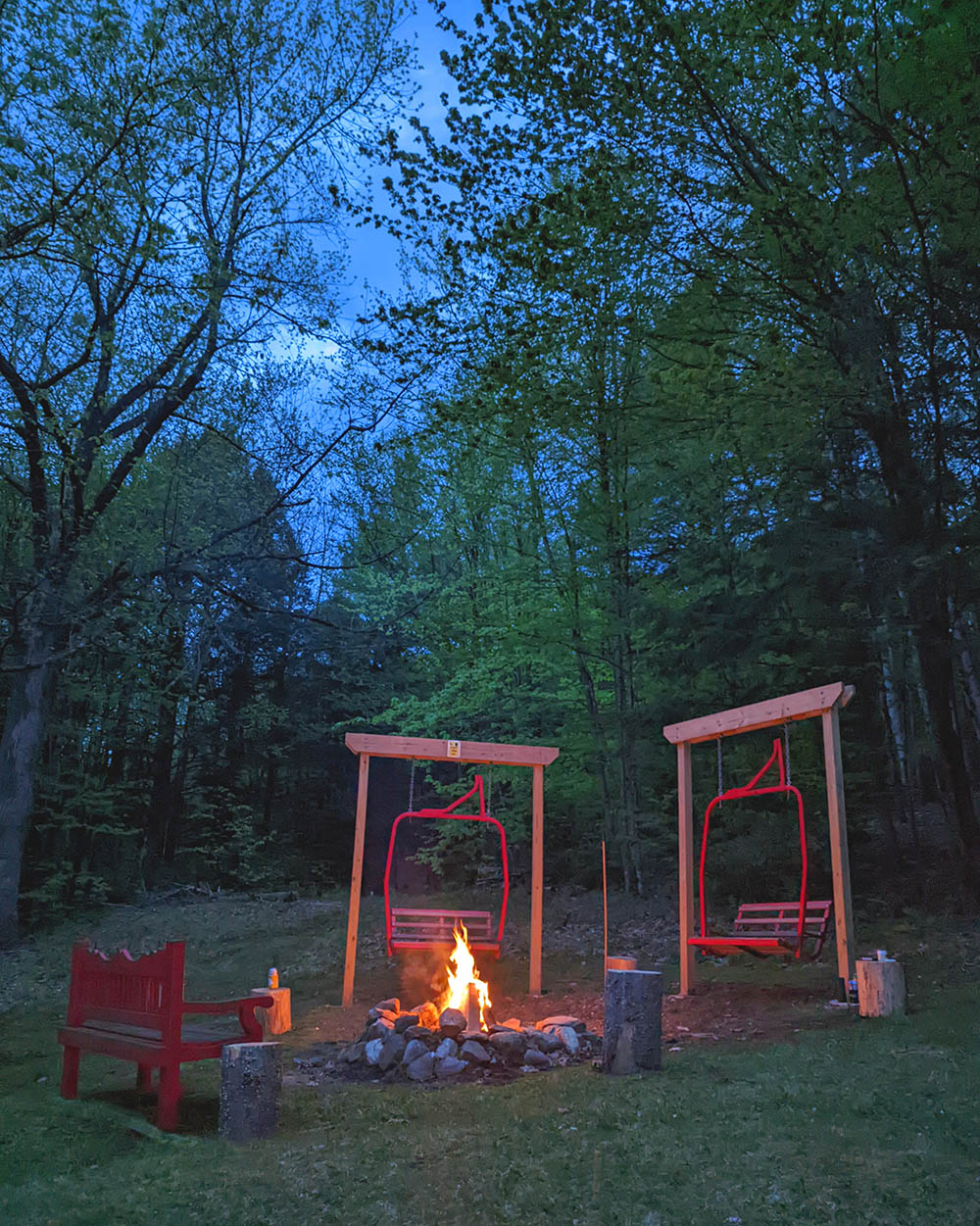 Two red swings are positioned beside a crackling campfire, surrounded by a wooded area. The fire flickers warmly, illuminating the space, while the soft twilight sky is visible through the trees. A cozy seating arrangement invites relaxation and connection.