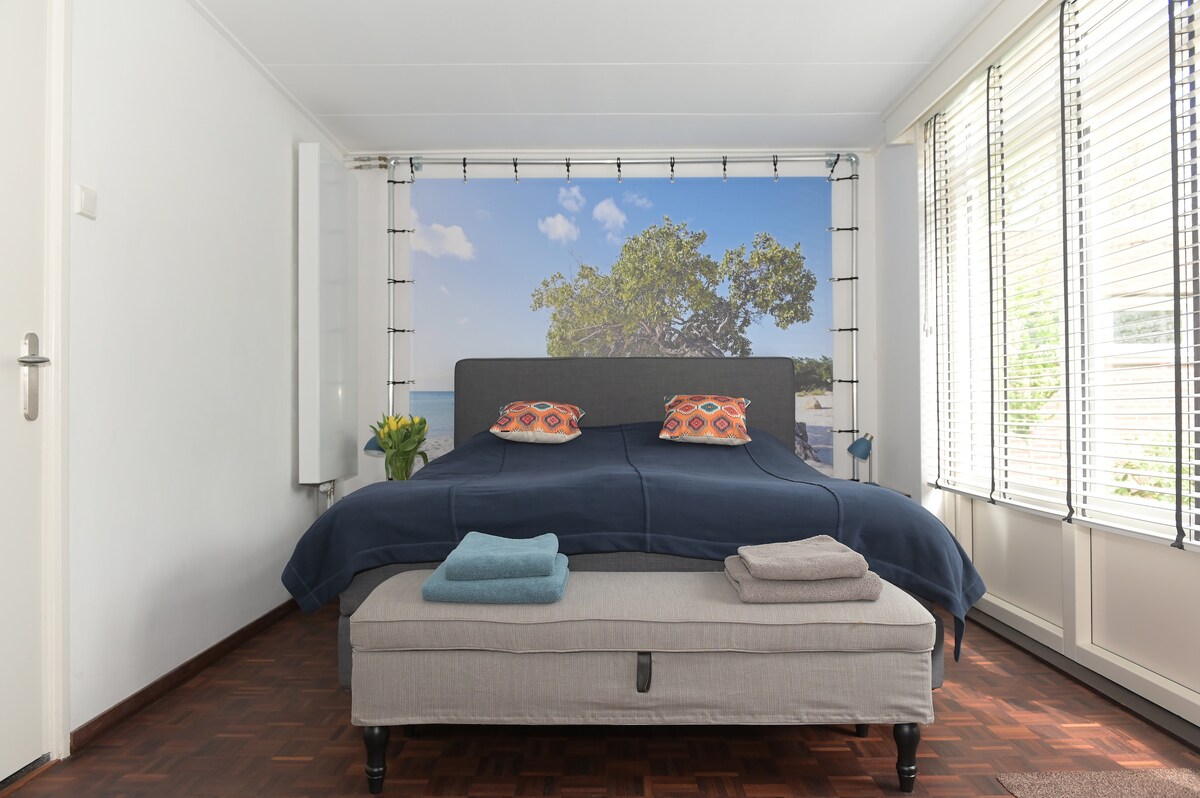 A spacious bedroom features a comfortable bed with decorative pillows. A bench holds neatly folded towels. Large windows allow natural light to fill the room, framed by sheer curtains. A nature-themed mural offers a calming backdrop, enhancing the overall serene environment.