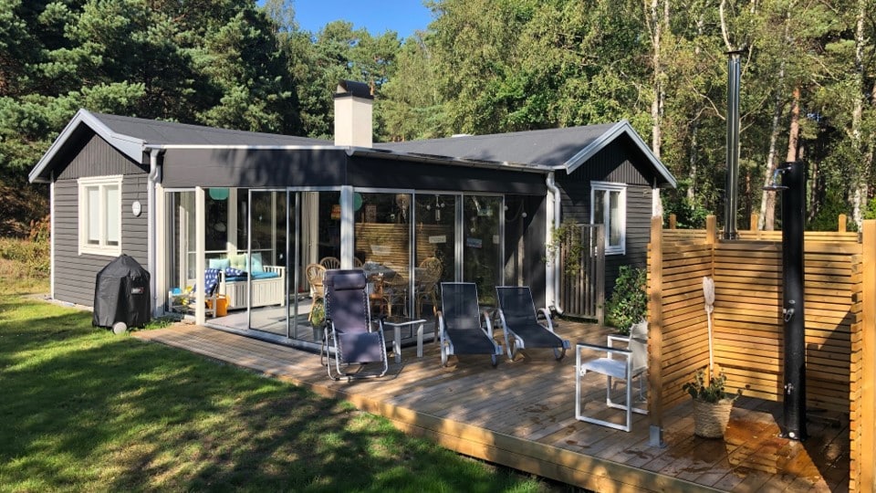 A modern cottage surrounded by trees features large glass doors that provide ample natural light. A spacious deck includes lounge chairs and an outdoor shower, enhancing the connection to nature. A wooden partition adds privacy while a grill is positioned nearby.