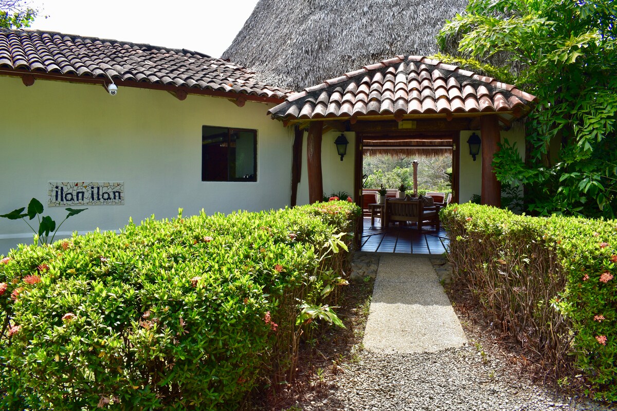 An inviting entrance path leads through lush greenery, flanked by flowering shrubs, toward a traditional bungalow featuring a thatched roof. The doorway offers a peek into a shaded outdoor dining area, creating a seamless transition between the vibrant garden and the cozy interior.