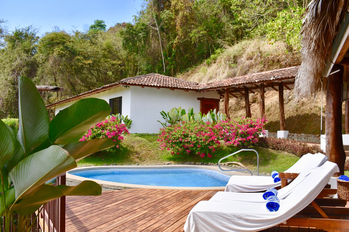 A private pool is set within a wooden deck, surrounded by lush greenery and vibrant flowers. The villa's white exterior is visible in the background, providing a sense of tranquility and connection to nature.