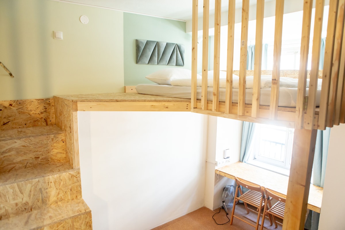 A compact private room designed like a Tiny House features a lofted sleeping area above a private bathroom. A wooden staircase leads to the sleeping space, while a table and chairs for work are positioned beneath the loft. Natural light enters from the nearby window.