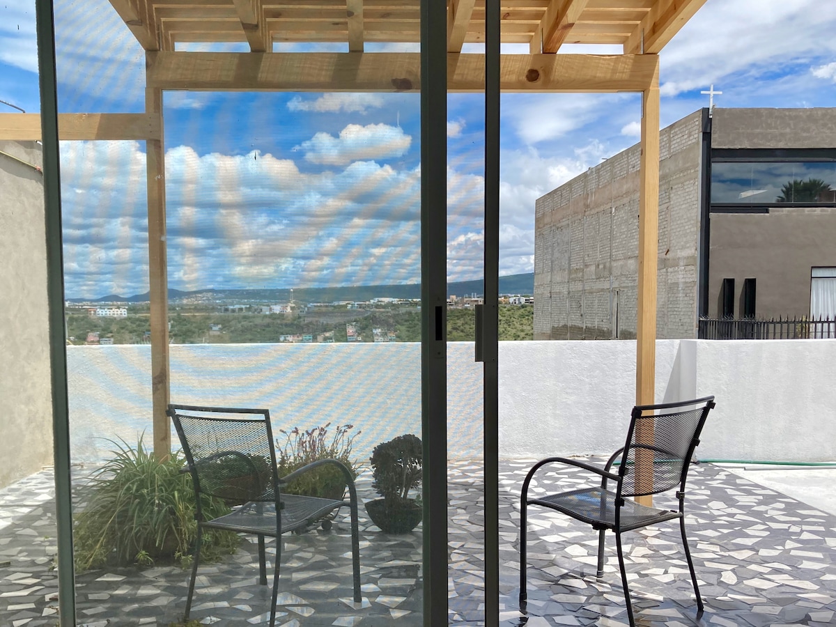 An inviting outdoor space features two metal chairs positioned on a stone patio. A wooden pergola provides shade, framing a view of the distant landscape filled with greenery and clouds. Potted plants enhance the natural ambiance.