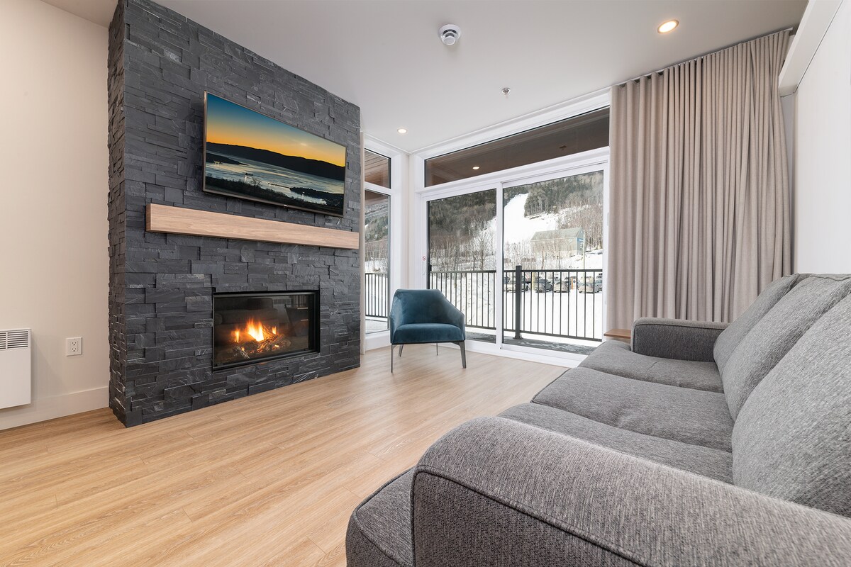 A spacious living area features a modern stone fireplace beneath a mounted television. Large glass doors provide access to a balcony with views of the surrounding landscape. Natural light fills the room, complemented by neutral-colored furniture and subtle curtains.