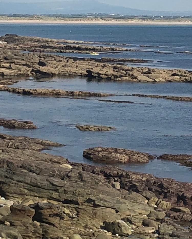 The rocky shoreline extends towards the calm waters of the Atlantic Ocean, showcasing various textures and colors of stones. In the distance, a sandy beach is visible, complemented by gentle waves lapping at the rocks.