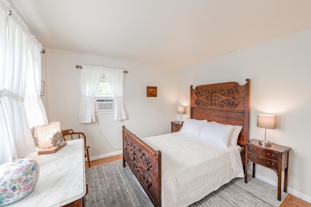 A tranquil bedroom is furnished with an antique wooden bed, complemented by matching nightstands and lamps. Large windows allow natural light to fill the space, while soft white curtains frame the view. A marble top dresser and a simple chair add charm to the room.